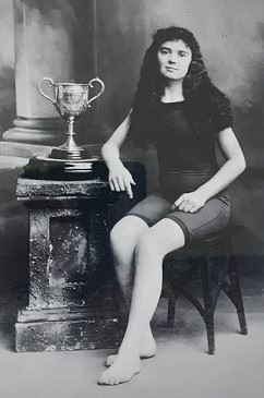 Black and white studio photo of young woman sitting on a chair beside a large silver trophy on a pedestal. She's wearing an old fashioned modest bathing suit, covering part of her upper arms and thighs. She has long black curly hair which is loose around her shoulders.
